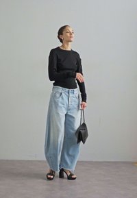 Cămașă neagră cu mâneci lungi, blugi deschiși la culoare, cu talie înaltă și cu picioare largi, sandale negre cu toc, ținând o poșetă mică neagră într-un cadru neutru.
