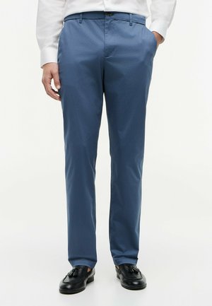 Person trägt blaue maßgeschneiderte Hose, weißes Hemd und schwarze Tassel-Loafers, steht mit einer Hand in der Tasche vor weißem Hintergrund.