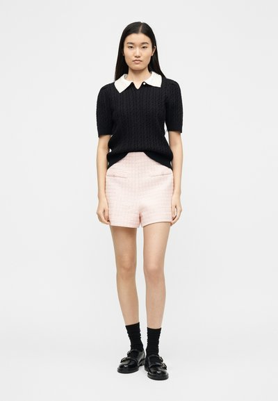 Jeune femme debout portant un pull à manches courtes noir avec col blanc, un short rose texturé, des chaussettes noires et des mocassins noirs sur fond blanc.