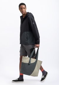 Lichte, zwart en donkergrijze hoodie-jack, gecombineerd met grijze shorts. Beige en grijze schoudertas met hengsels. Zwarte schoenen met witte zolen.