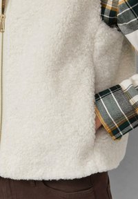 Weißer, flauschiger Fleece-Weste mit einem goldenen Reißverschluss, kombiniert mit einem grün-gelben Karoshirt, das an den Manschetten sichtbar ist. Texturierte Oberflächendetails.