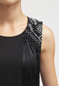 Vestido negro con intrincados bordados blancos en el hombro. Presenta flecos largos y detallados en el costado, creando textura y movimiento.
