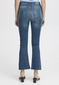 Jeans in denim a campana di colore blu scuro con vita aderente, due tasche posteriori e bordi grezzi all'orlo. Indossati con sandali neri con tacco a blocco.