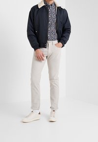 Granatowa kurtka bomberka z kołnierzem z kożucha, kwiecista koszula na guziki, jasnobeżowe spodnie i kremowe trampki. Codzienny strój z różnorodnymi fakturami.