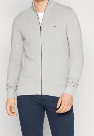Mann trägt hellgrauen Zip-Pullover mit kleinem Logo auf der Brust und dunkelblaue Hose, steht vor einfarbigem Hintergrund.
