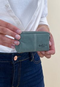 Zelená kožená peňaženka s bielym prešívaním, obdĺžnikového tvaru, s uzáverom na zips a embosovaným logom na prednej strane.