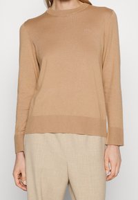 Femme portant un pull à manches longues beige avec un petit logo « BOSS » sur la poitrine et un pantalon beige clair.