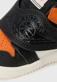 Baskets en cuir noir avec des accents orange, présentant un dessus texturé et un logo embossé. Comprend une semelle intercalaire blanche et un design de pointe perforé.