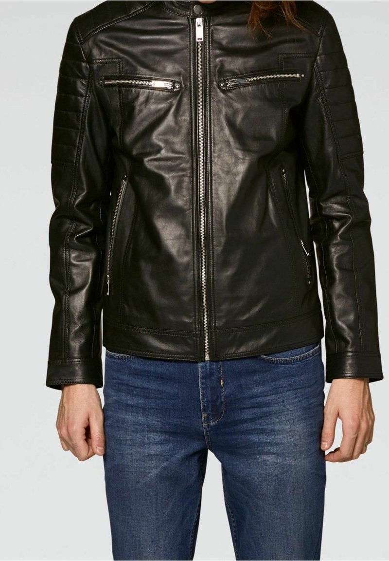 Veste en cuir noire à coupe ajustée, avec deux poches poitrine zippées et des détails côtelés sur les épaules. Portée avec un jean bleu.