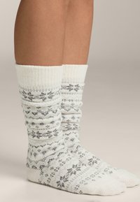 Bela pletena nogavica s sivimi vzorci, z rebrastim robom in zbrano teksturo, ki poudarja udoben in topel videz.