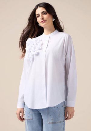 Camicia bianca con bottoni, caratterizzata da un motivo floreale in rilievo sulla spalla sinistra. Vestibilità ampia, maniche lunghe e tessuto di cotone morbido.
