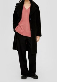 Manteau noir et confortable avec un col châle, deux boutons, porté sur un pull rose à col en V. Le modèle l'associe à un pantalon noir ample et des chaussures noires épaisses.