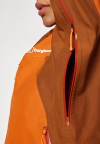 Orange and brown jacket with a zippered pocket, featuring a drawcord and branding. Material is smooth with a slight sheen.