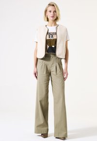 Gilet en polaire beige porté sur un t-shirt graphique blanc avec un accent léopard, associé à un pantalon large vert olive et des talons à motifs.