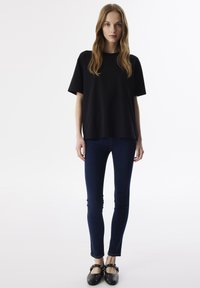 Zwarte korte mouwen t-shirt, blauwe skinny jeans en zwarte platte schoenen met studs. De stof lijkt soepel; de pasvorm is relaxed en casual.