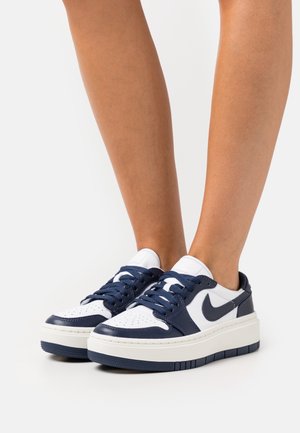 Person wearing low-cut white and navy blue Nike sneakers with thick soles and navy laces, standing on a plain white surface.