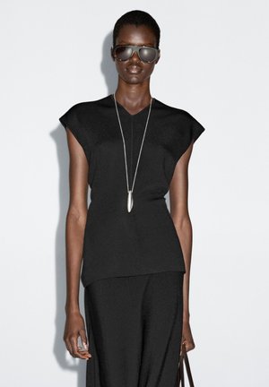 Top nero a maniche corte con scollo a V e design sartoriale, abbinato a una lunga gonna nera. La modella indossa occhiali da sole scuri e una lunga collana d'argento.