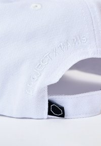 Gorra de béisbol blanca con tejido texturizado, bordado "PERFECT PARIS" y una etiqueta negra con un emblema geométrico en la parte posterior.