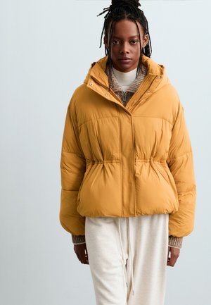 Modèle portant une doudoune à capuche jaune moutarde par-dessus un pull en maille et un pantalon crème, debout devant un fond clair.