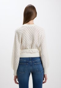 Maglione bianco lavorato a maglia con texture a motivo diamante, maniche a palloncino lunghe e dettaglio in vita arricciato. Abbinato a jeans in denim blu.
