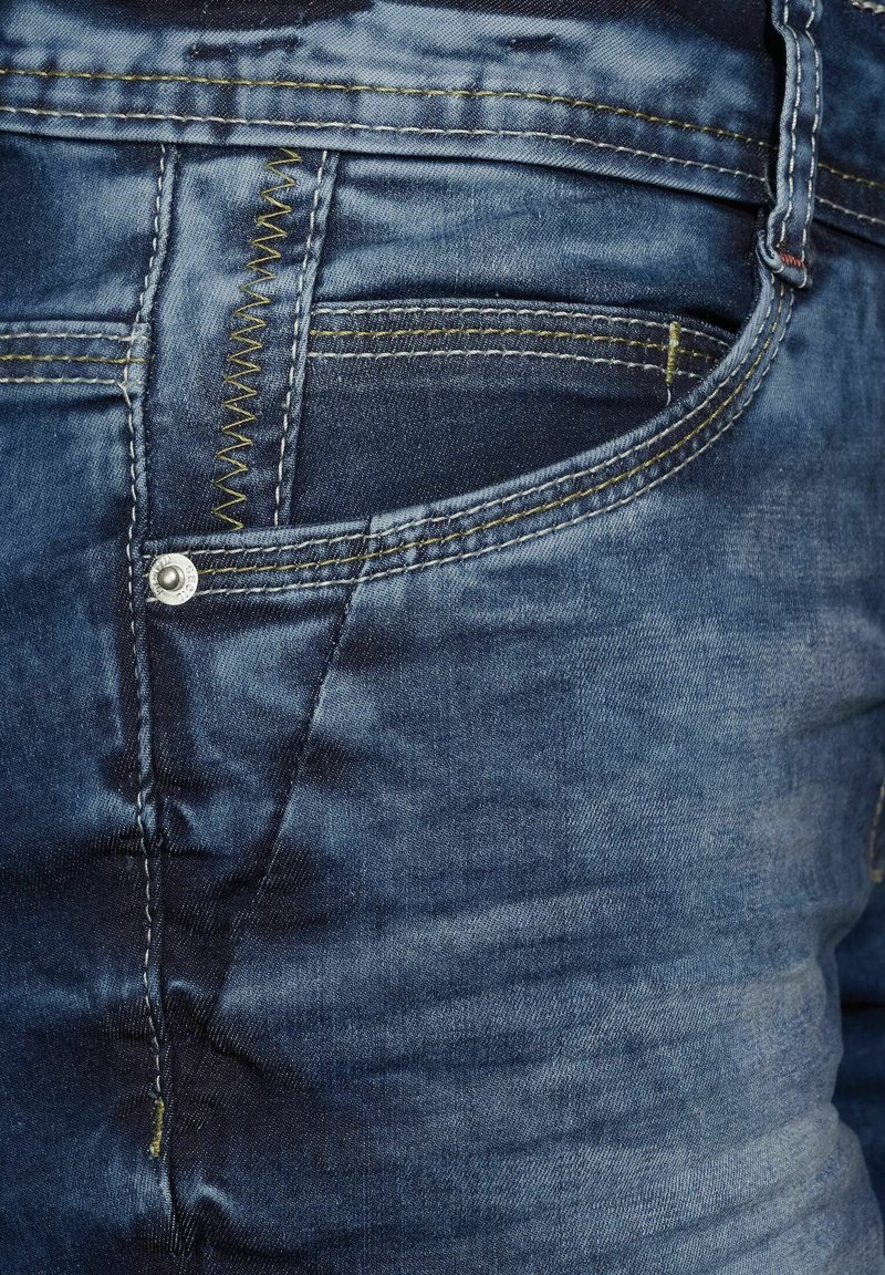 Gros plan sur un jean en denim bleu montrant la poche avant, le rivet et les détails de couture jaune sur un tissu texturé.