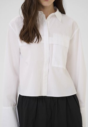 Cămașă albă, cu butoane, scurtă, cu mâneci lungi, având un buzunar frontal și o textură netedă. Combinate cu o fustă neagră, pliată.