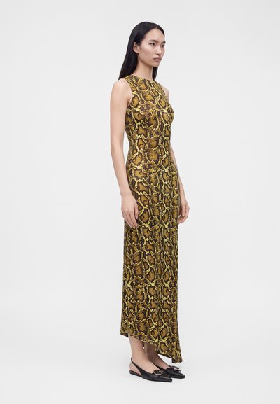 Femme aux longs cheveux noirs portant une robe maxi sans manches jaune et marron à imprimé serpent et des ballerines slingback noires, debout devant un fond uni.