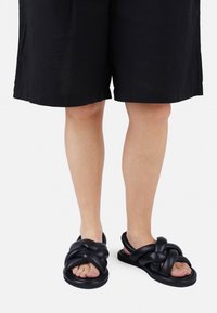 Sandalias negras con un diseño anudado, hechas de material suave. Cuentan con la punta abierta y una suela gruesa y redondeada.