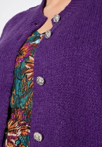 Lilas gestrickte Strickjacke mit einer strukturierten Oberfläche, versehen mit zwei metallischen Löwenknöpfen und einer bunten gemusterten Bluse darunter.