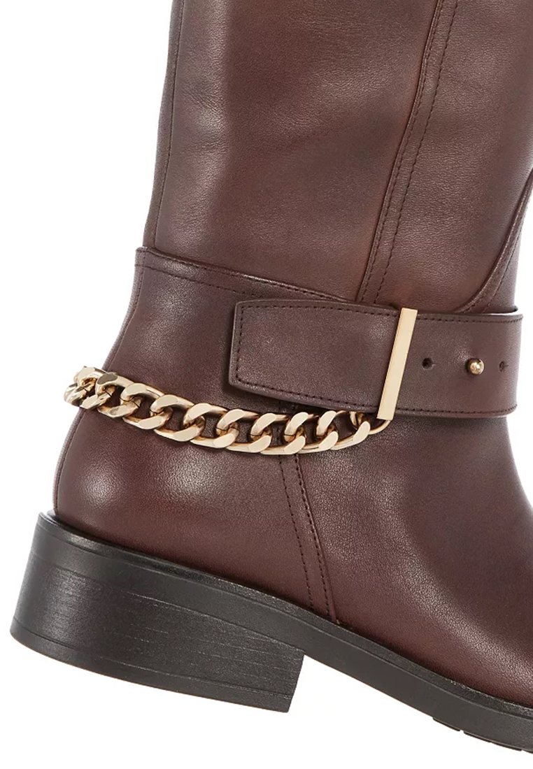 Stivale in pelle marrone con punta rotonda, tacco basso a blocco, con un accento in catena dorata attorno alla caviglia e un dettaglio con fibbia.