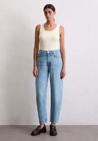 Lys gul tanktop, lys blå high-waisted jeans og mørkebrune loafers. Myk stoff, avslappet passform, lett bleking på jeans.