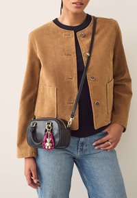 Bruine suède jas met ronde halslijn en knoopsluiting aan de voorkant, gecombineerd met een kleine zwarte handtas met een roze decoratieve hanger.