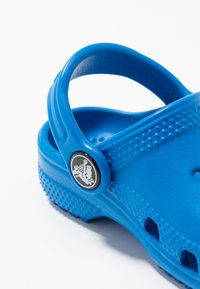 Detailní záběr modrého gumového clogu s texturovanou podešví a nastavitelným páskem na patě, který má černé kulaté logo na knoflíku.
