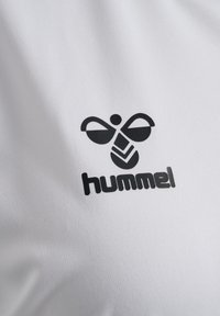 Biały materiał o gładkiej fakturze z czarnym logo przedstawiającym stylizowaną pszczołę oraz napisem "hummel" pogrubioną czcionką.