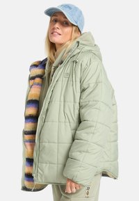 Lichtgroene gewatteerde pufferjack met kleurrijke gestreepte voering, gecombineerd met een grijze hoodie en een lichtblauwe spijkermuts.