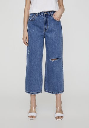 Korkeavyötäiset, leveälahkeiset farkut, jotka on valmistettu sinisestä denimistä ja joissa on kulunut ilme. Farkuissa on iso repeämä reidessä ja räjähtäneet lahkeet.