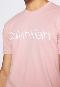 Różowa bawełniana koszulka z okrągłym dekoltem i krótkimi rękawami, z napisem "Calvin Klein" wydrukowanym na białym tle na piersi.