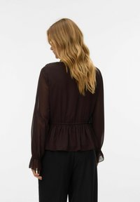 Blouse longue à manches longues couleur bordeaux profond avec une taille peplum, tissu doux, silhouette fluide et poignets froncés. Détail de boutons au dos.