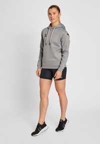 Haut à capuche gris avec cordons, accents de logo noirs, associé à des shorts noirs et des chaussures de sport noires, se tenant sur un fond uni.