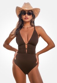 Brązowy, prążkowany jednoczęściowy strój kąpielowy z głębokim dekoltem w kształcie V, wcięciem w talii oraz złotymi akcentami. W połączeniu z kapeluszem ze słomy o szerokim rondzie i okularami przeciwsłonecznymi.