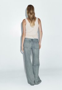 Rozšířené modré džíny s bledým odstínem a dvojitými zadními kapsami, v kombinaci s krémovým tílkem, pohled zezadu.