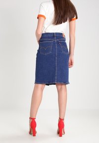 Denim pencil skirt in dark blue with a fitted waist, two back pockets, and a raw hem. Complemented by bright red high-heeled shoes.