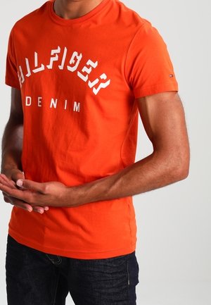 Mężczyzna ubrany w jaskrawo pomarańczową koszulkę Tommy Hilfiger Denim z białym logo, złożonymi rękami z przodu, na jednolitym jasnym tle.