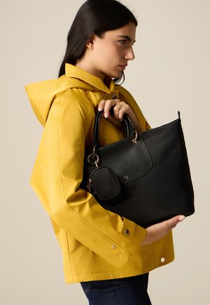 Giovane donna con giacca a vento gialla che tiene una borsa nera con una piccola pochette su uno sfondo uniforme.