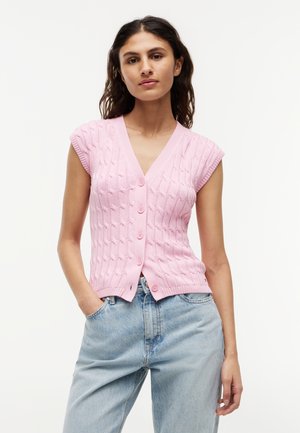 Žena s dlhými tmavými vlasmi, oblečená v ružovej pletenej veste bez rukávov so zapínaním na gombíky a svetlomodrých rifliach, stojaca s jednou rukou v vrecku.