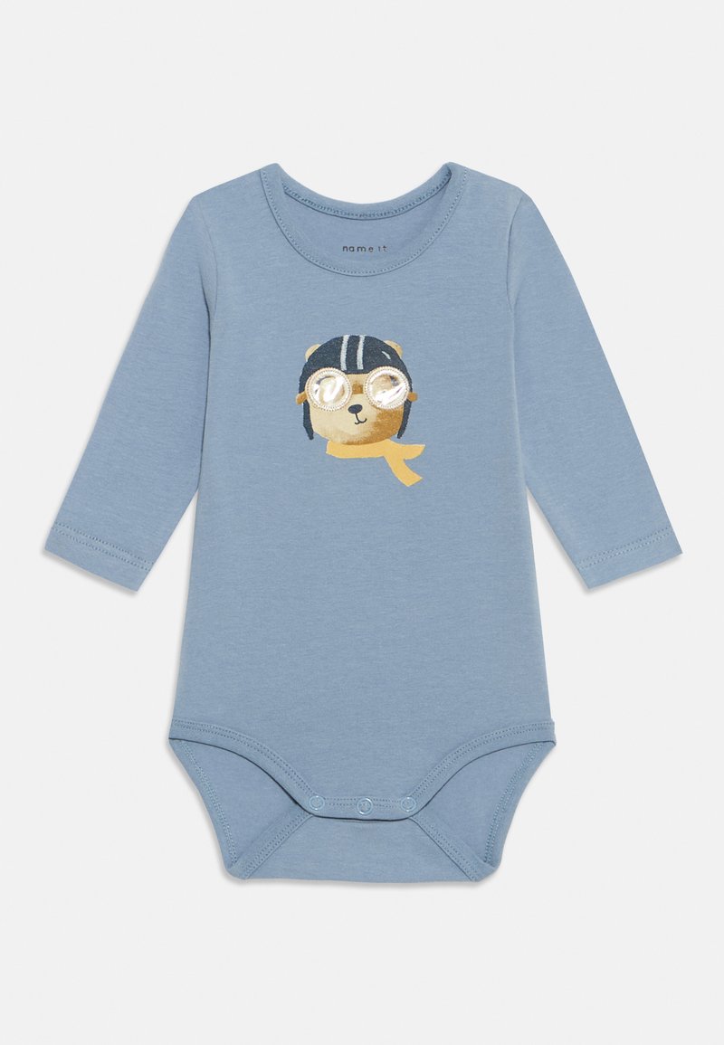 Šviesiai mėlyna ilgomis rankovėmis kūdikio kombinezonas su spausdinta meškos, dėvinčios akinius ir šalį, dizainu. Apatinėje dalyje yra spaudės užsegimai.