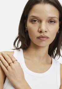 Goldfarbene Metallohrringe und Ringe mit glatten, geschwungenen Formen. Modell trägt ein weißes Tanktop mit einem runden Ausschnitt.