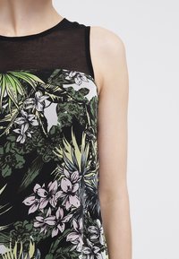 Dámská bezrukávová blůza s květinovým potiskem a černým průhledným výstřihem, živé zelené a růžové vzory na tmavém pozadí, hladká textura materiálu.