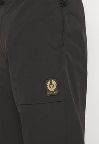 Černé cargo kalhoty s texturovaným povrchem, na stehně mají zlatou nášivku Belstaff a boční kapsu se zipem.