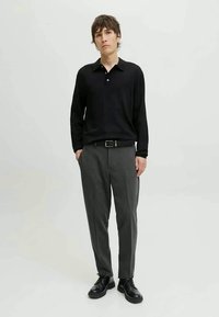 Camisa polo preta de manga longa com colarinho, combinada com calças cinzentas ajustadas. Sapatos de couro pretos completam o look. Fundo simples.